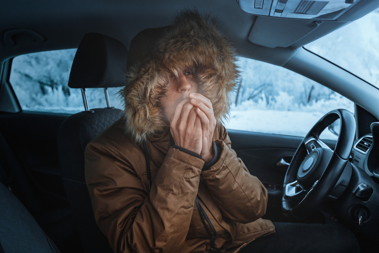 A man sits in a freezing cold car with his hood up, blowing on his hands. 